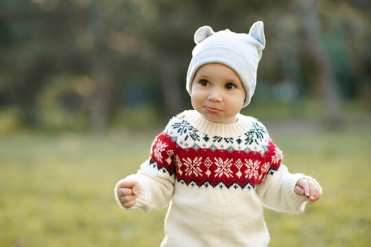 Smiling kid boy 1 year old wearing knitted xmas sweater and hat walking in autumn park. Childhood. Looking at camera