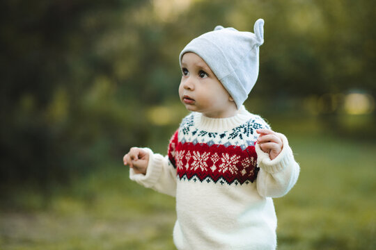 Cute kid boy 1 year old wearing knitted xmas sweater and hat walking in autumn park. Childhood. Looking away.