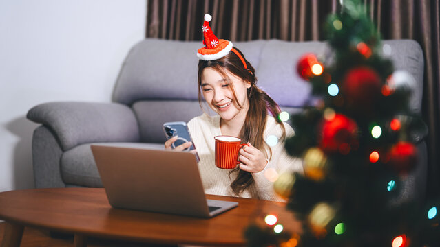 Young adult asian woman using laptop relax sitting in cozy winter living room with christmas tree enjoy alone at home for the holidays