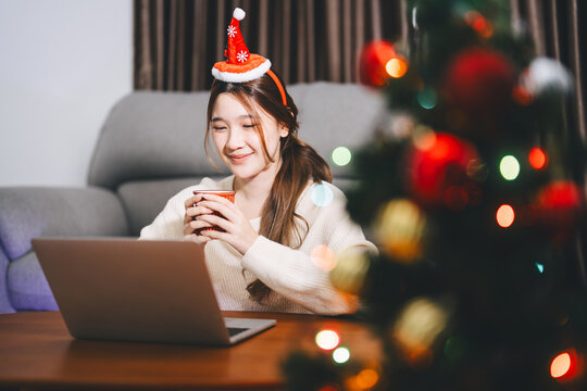 Young adult asian woman using laptop relax sitting in cozy winter living room with christmas tree enjoy alone at home for the holidays