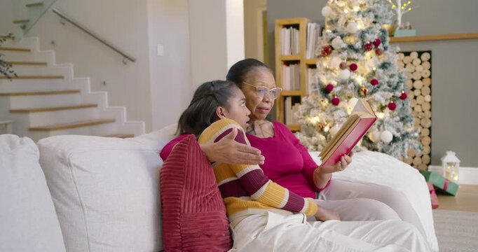 African American senior woman and youth girl opening book, reading, sharing on sofa by lit tree