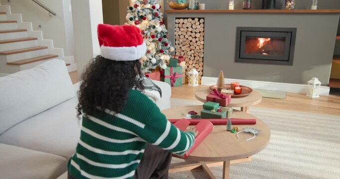 African American woman wearing Santa hat placing box on red paper, wrapping at home preparing gifts
