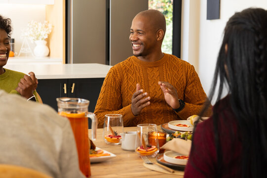 Diverse friends sitting at table in kitchen, man in rust orange sweater smiling, pitcher near