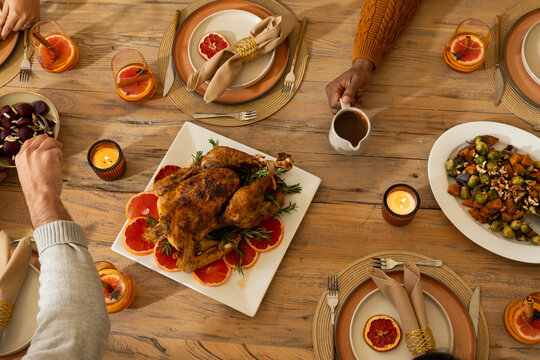 Diverse friends passing dishes and pouring gravy at wooden dining table with roasted chicken
