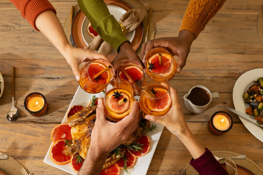Five friends wearing cozy sweaters clinking cocktails at dining table with roasted poultry