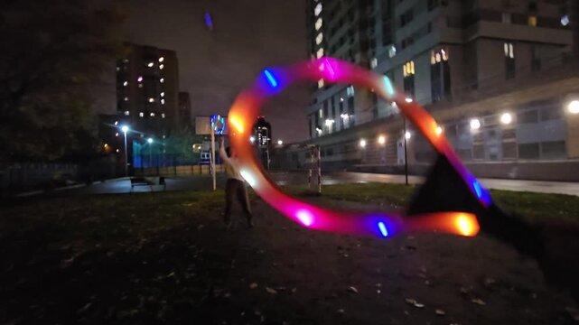 Nighttime city park with glowing LED hula hoop. Person holds hoop, lights flash in motion. Background shows city buildings and streetlights. Dark grass and pavement under illuminated scene