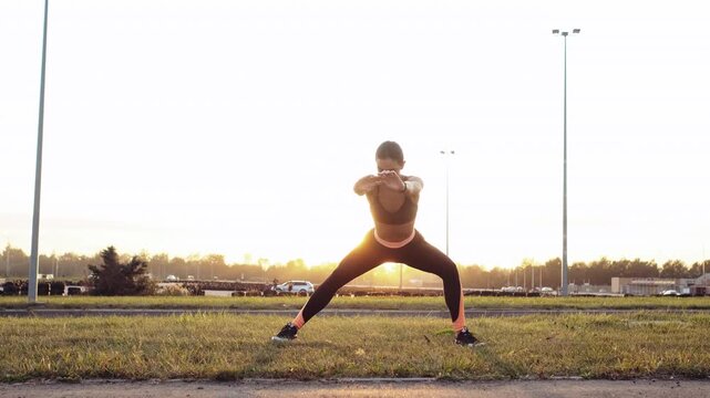 Young woman holding controlled wide lateral lunge on grassy strip beside roadway at sunrise or sunset, weight on bent leg with opposite leg extended and arms crossed at chest for balance, focused