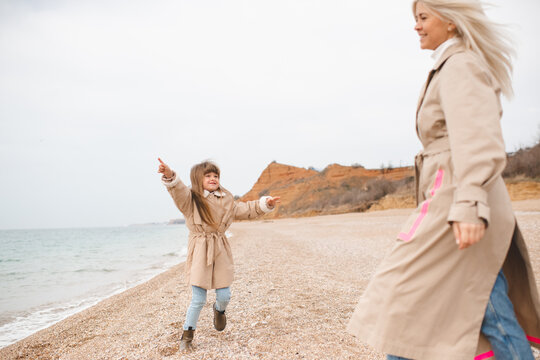 Child girl having fun with her mother wearing similar beige jackets at beach over sea. Childhood. Autumn season.
