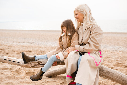 Mother holding child daughter sitting at log at beach wearing similar beige coats spending time together. Motherhood. Autumn season.