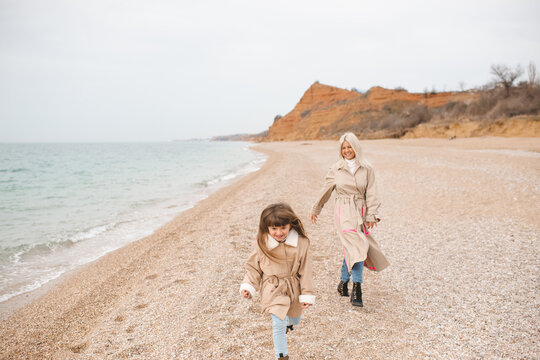 Happy child girl running with her mother at beach wearing beige jackets at sandy beach together. Autumn season.