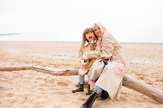 Mother holding child daughter sitting at log at beach wearing similar beige coats spending time together. Motherhood. Autumn season.