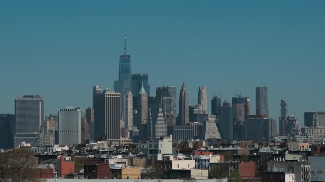 Frontal panoramic motion scene showing dense cluster of highrise office and residential towers rising above low-rise brick and masonry rooftop neighborhood, above-ground subway train occupies lower