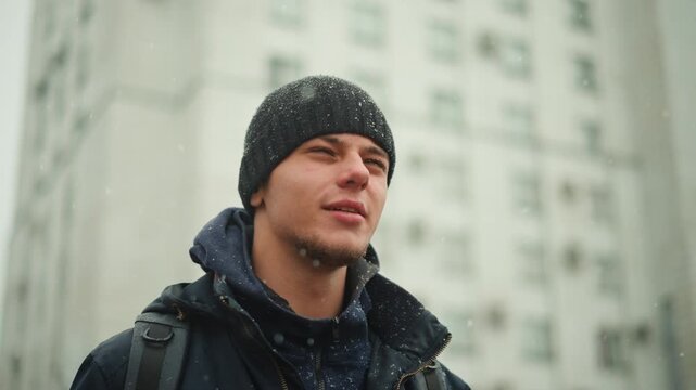 Close up of man wearing winter clothing with snow falling around him. subject is dressed in dark outerwear including jacket and beanie while carrying backpack. Tall buildings are visible