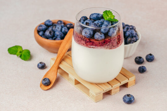 Creamy yogurt dessert topped with fresh blueberries and mint leaves served in a glass on a wooden coaster with bowls of blueberries in the background