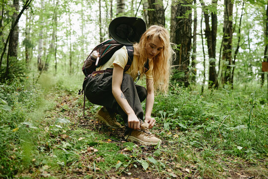 Young Caucasian woman hiker crouching to tie shoelaces on forest trail during outdoor trekking adventure. Image ideal for active lifestyle marketing and ecotourism campaigns, eye level shot