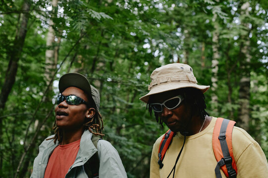 Two young adult African American men hiking in green forest, wearing hats and sunglasses. Image suitable for outdoor recreation campaigns and active lifestyle marketing, medium shot