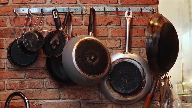 Pans hanging on brick kitchen wall, cooking utensils display. Metal cookware hangs from hooks, creating a rustic storage and home interior design concept.