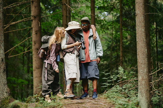 Diverse young adult friends hiking in forest, looking at camera screen together. Image represents outdoor recreation and is suitable for travel and adventure marketing campaigns