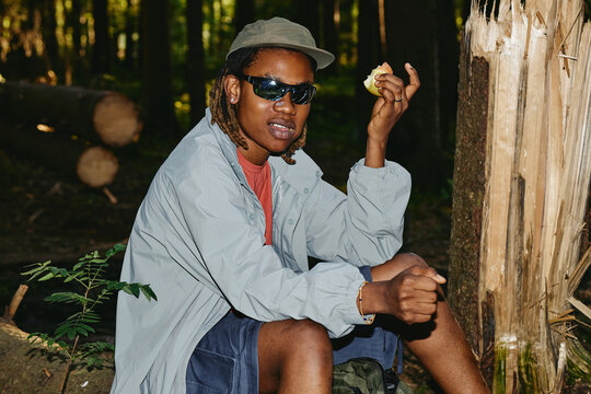 Portrait of young Black man sitting in forest during hike and holding apple. Image represents outdoor recreation and healthy lifestyle, suitable for eco tourism campaigns