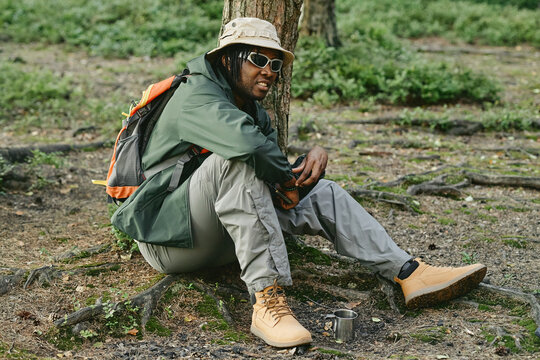 Young adult African American man resting by tree during forest hike, promoting outdoor recreation and wellness. Image suitable for active lifestyle marketing campaigns