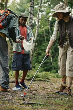 Vertical image of diverse young adult volunteers collecting plastic litter in forest using trash grabber tool, promoting environmental conservation and community cleanup campaigns