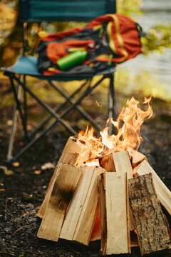 Vertical shot of campfire burning in forest with camping chair and backpack in background, representing outdoor adventure. Concept of active recreation