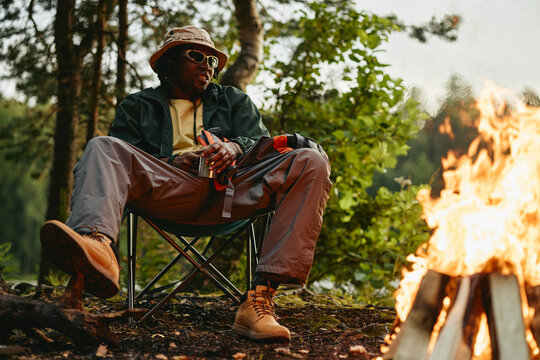 Young adult African American man sitting on folding chair near campfire in forest during hiking trip. Image suitable for outdoor gear advertising and adventure travel promotions