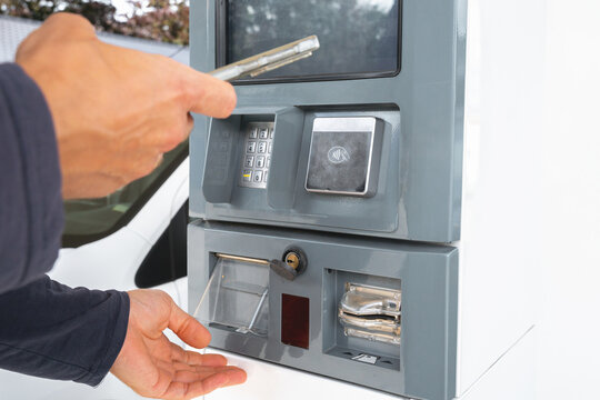 Close-up of hands using a smartphone for contactless payment at an outdoor service terminal. The grey machine includes a keypad and card reader near a vehicle.
