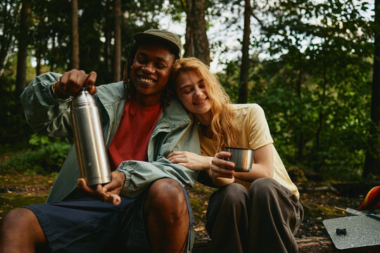 Diverse young adult couple sitting in forest during hiking trip, African American man holding thermos and Caucasian woman smiling. Image suitable for outdoor recreation marketing