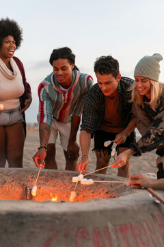 Group of friends roasting marshmallows around a beach fire pit.