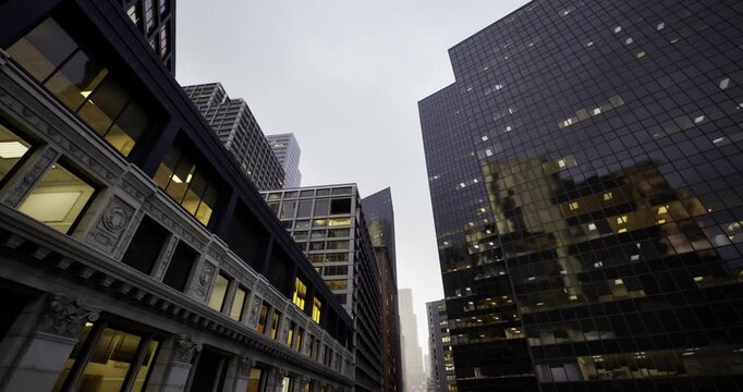 moody passage between reflective highrises with warm interior lights visible through windows, strong vertical lines and converging perspective creating