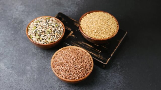 Wooden bowls filled with mixed seeds, sesame seeds, and flax seeds arranged on a dark background