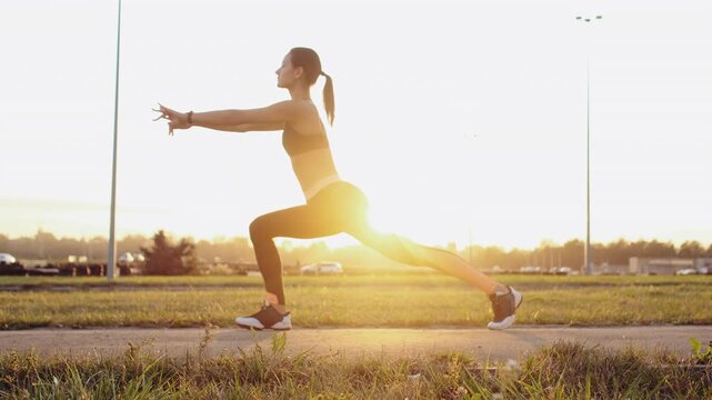 Profile of young woman in dark sports bra and black leggings holding forward lunge on paved path beside grassy verge at sunrise, with woman, lunge, exercise, fitness detail, showing athlete, workout