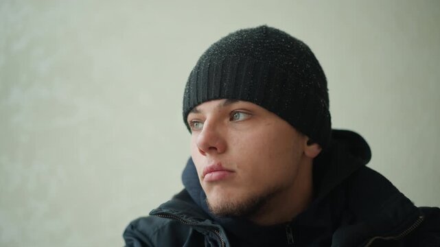 Close up of man wearing black beanie and dark jacket seated against plain light colored background. Subject remains stationary with subtle head movements. Soft lighting creates even