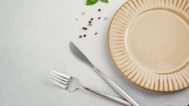 A beige plate with fork and knife on a light grey background with peppercorns and basil leaves