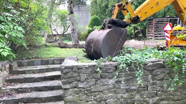 A yellow excavator arm rests above a trench near old stone stairs outdoors, site preparation, landscaping process, construction development.