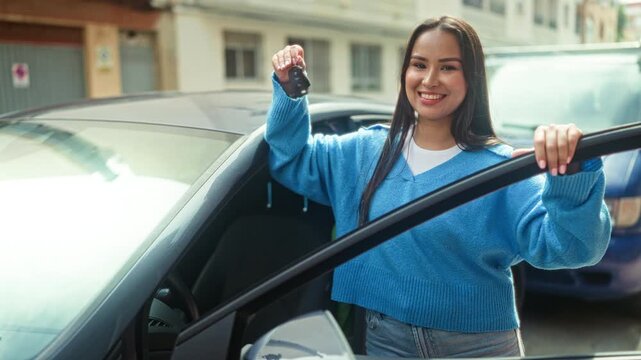 Woman young latin hispanic smiling, holding car key in right hand and pointing at open car door while standing on a street beside parked cars; joyful independence.