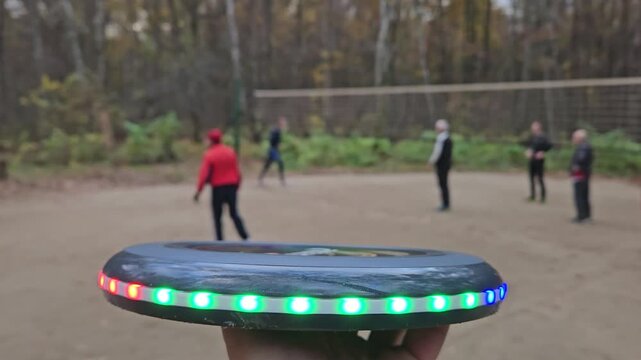 Hand holds glowing disc with LED lights. People play flying disc plate in wooded park area. Disc features alternating red, green, and blue lights. Lighting enhances visibility during outdoor activity
