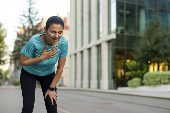 Young woman runner clutching chest and knee, gasping for breath on a city street, struggling with sudden chest pain and respiratory distress during exercise