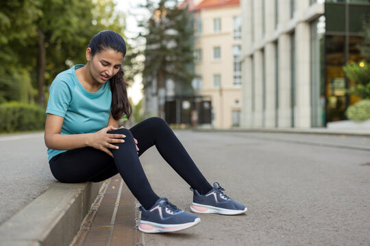Young woman sitting on a curb, holding her injured knee with a grimace of pain, interrupting her run on an urban street due to a sports injury or discomfort