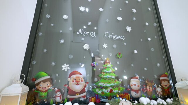 Glass window decorated with Christmas stickers. Santa and elves surround a miniature tree. Cute cat walks past the display. Warm lanterns glow beside the window