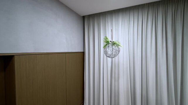Silver wreath hangs in modern, softly lit living room. Foliage and pinecone adorn the decorative sphere. White curtains frame the stylish interior. Background reveals a tree and cozy furnishings