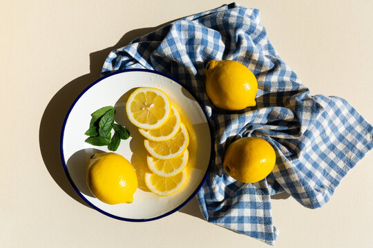 Rondelles de citrons jaunes, un citron entier et feuilles de menthe pos&eacute;s sur une assiette blanche &eacute;maill&eacute;e avec un liser&eacute; bleu, torchon &agrave; carreaux bleus pos&eacute; &agrave; c&ocirc;t&eacute; avec deux citrons, vue du dessus
