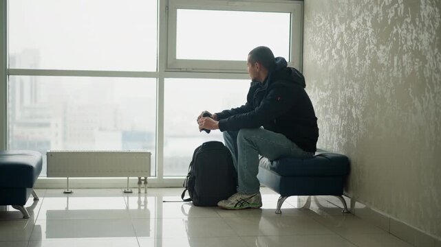Close up of man sitting on bench near large window with city view holding object in hands. Black backpack placed beside him soft natural light illuminates indoor space featuring tiled floor