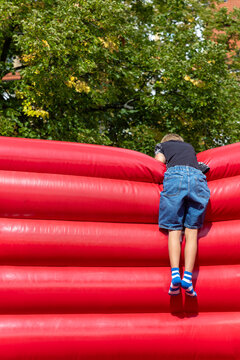 Child climbs red inflatable obstacle outdoors, viewed from behind against green trees in sunny park. Active summer play captures adventure, balance, effort, fun, childhood, recreation and energy