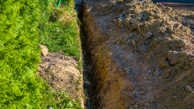 Long, narrow trench freshly excavated in residential backyard garden alongside dense green hedge, showing piles of loose brown soil during outdoor construction project
