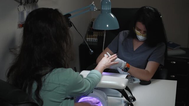 Manicure technician applies gel and spot fixes nail tip while client cures other hand under ultraviolet lamp during salon procedure