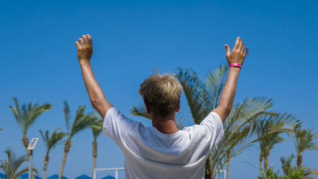 Young Man With Blonde Hair Standing With Arms Raised High Toward Bright Blue Summer Sky Against Backdrop Of Tropical Palm Trees On Sunny Vacation Day At Resort