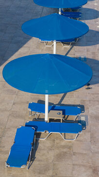 Aerial view of row of empty blue lounge chairs and large blue umbrellas set up on tiled patio, casting long shadows on sunny day, suggesting resort or pool area awaiting visitors