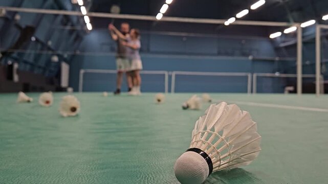 Badminton shuttlecocks lie scattered on the court floor. Two players stand behind the net in an indoor facility. Lighting fixtures illuminate the high ceiling. Shuttlecocks are white with black bands